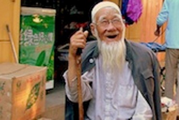 An old man, smiling with his walking stick sit on a stool